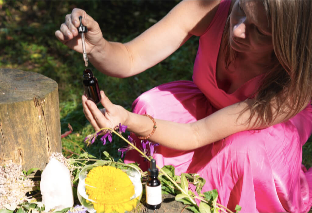 Untitled Blonde woman in a pink dress, holding an open bottle of flower essence in front of a log and flowers